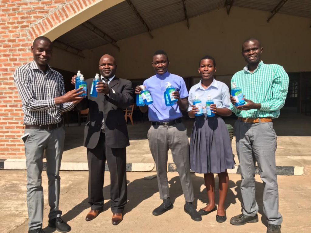 Headteacher in Black Suit and students in uniform receive cholera prevention materials fom Mtuli Foundation